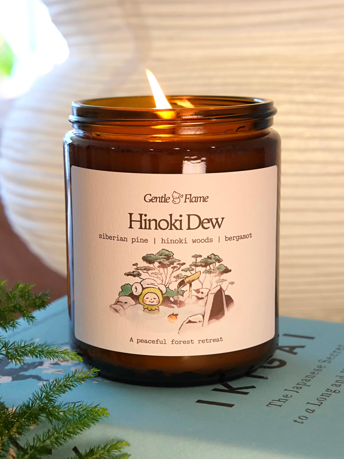 Candle in an amber jar labeled 'Hinoki Dew' by Gentle Flame, with a lit wick on a light background.